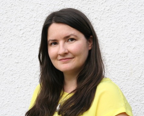 Photo of a woman with a warm smile and long brown hair wearing a yellow top and standing against a while background.